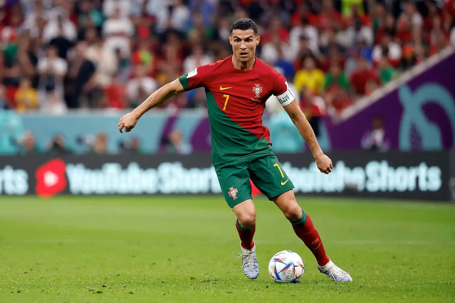 A number 7 soccer player in red and green dribbles the ball, with the crowd and ads in the background